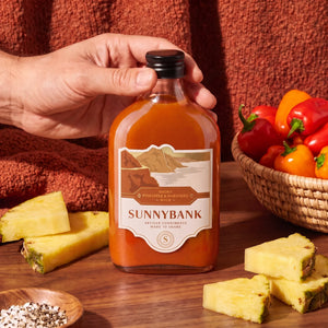 Hand holding a bottle of Sunnybank mild smoky pineapple and habanero hot sauce beside pineapple slices and peppers.