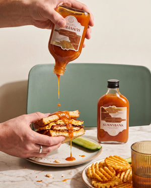 Sunnybank pineapple and habanero hot sauce being drizzled over a sandwich beside waffle fries.