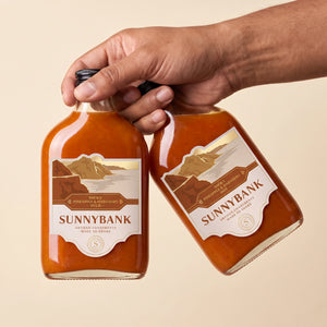 Two bottles of Sunnybank smoky pineapple and habanero hot sauce held against a beige background.