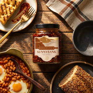 Jar of Sunnybank chilli crisp oil beside baked beans, fried egg and toast on a wooden breakfast table.