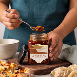 Spoonful of Sunnybank chilli crisp oil held above the jar beside pizza and dumplings.