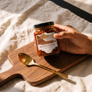 Hand holding a jar of Sunnybank chilli crisp oil over a wooden board beside a gold spoon.