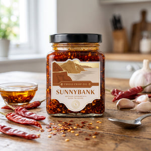 Jar of Sunnybank smoky pineapple chilli crisp oil with dried red chillies, garlic and a spoon on a wooden table.